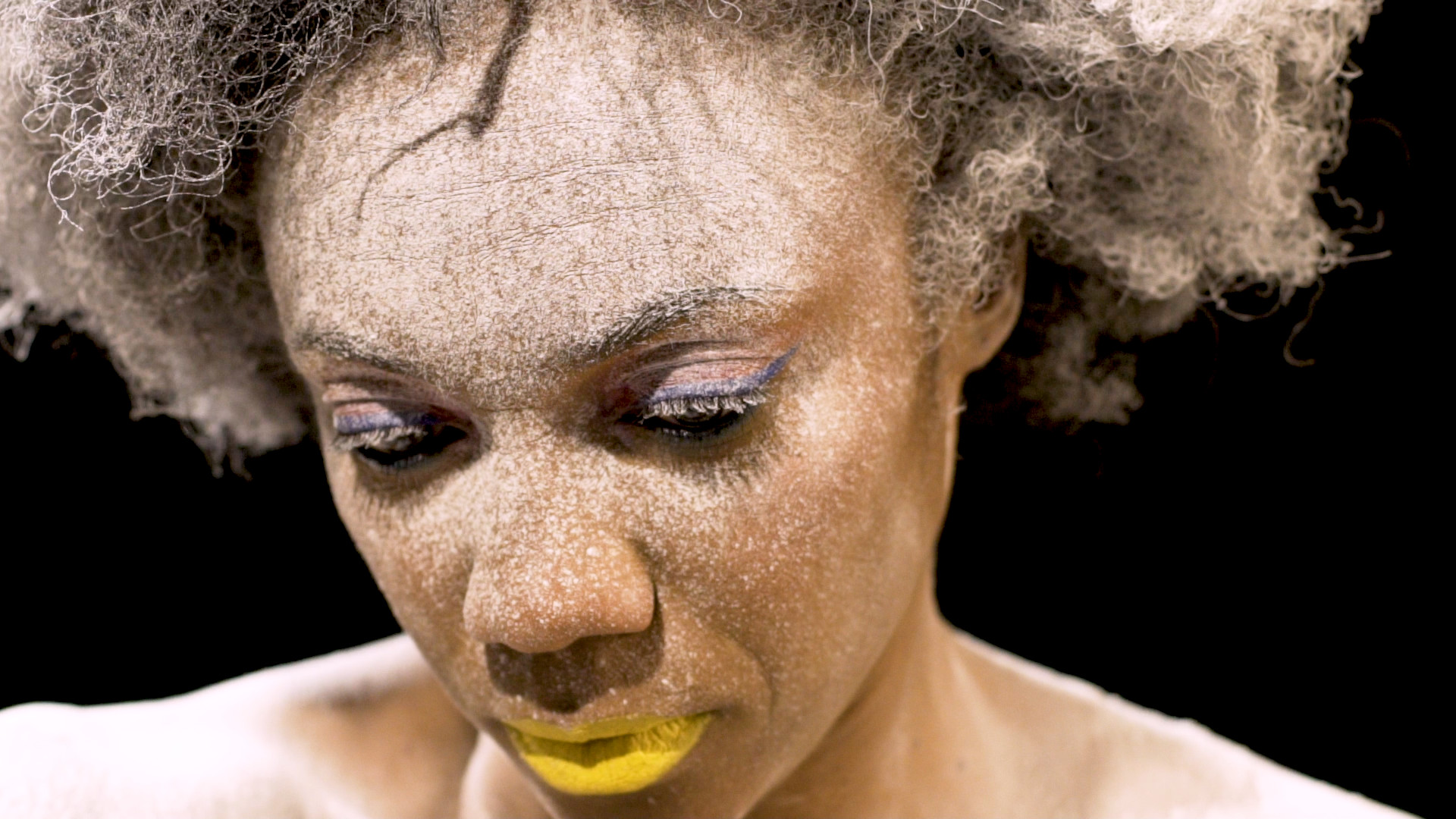 A woman with powder on her face and yellow-painted lips looks down mournfully.